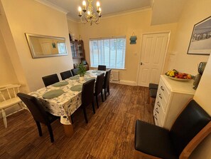 A dining area with a table set for six, surrounded by chairs, a chandelier, a window with blinds, and a cabinet with fruit. - Seaside Holiday Retreat in Filey, North Yorkshire 
4 Bedrooms - Sleeps 10 (Filey)