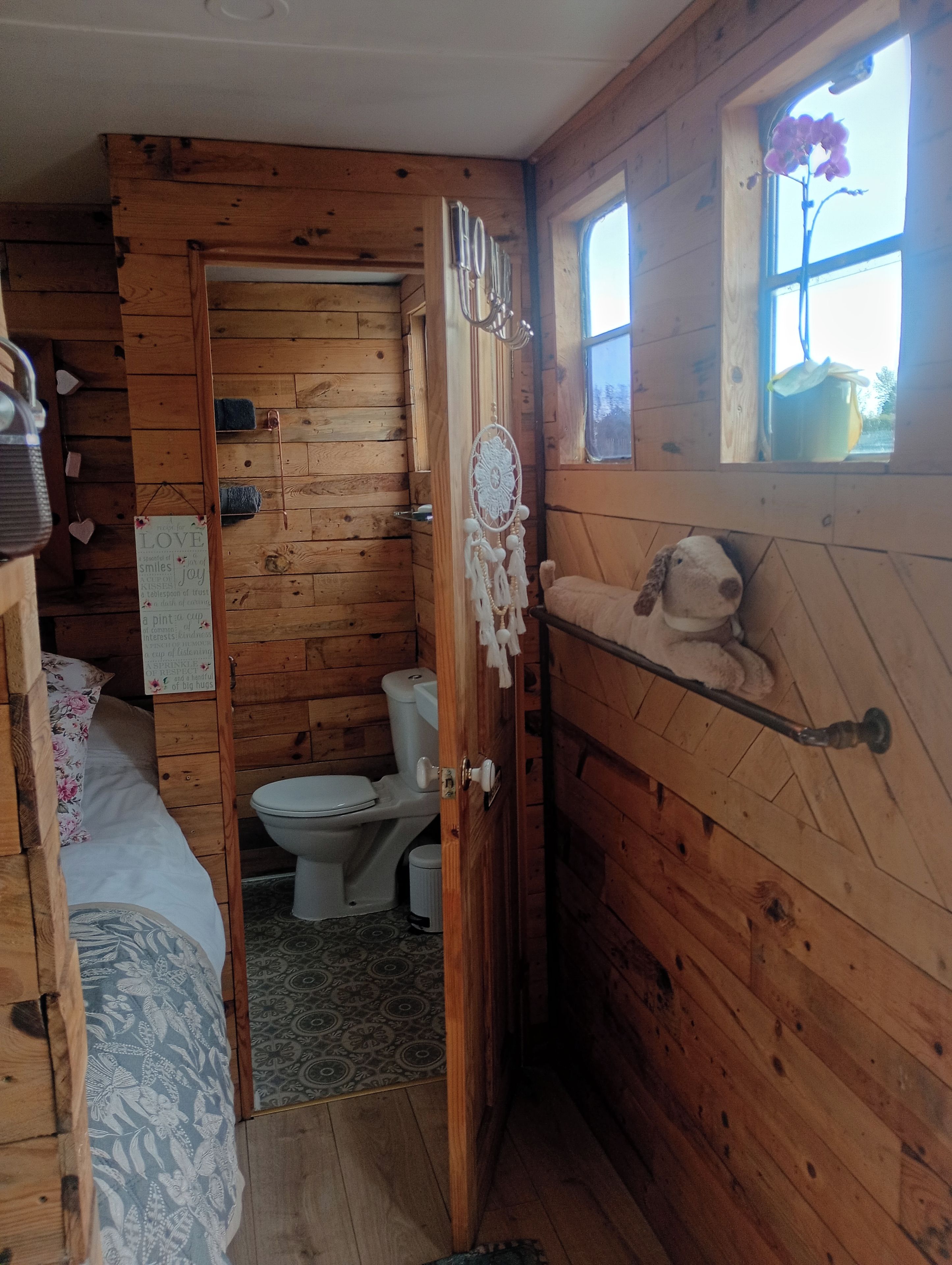 Puinen mökki sisältä, siellä on sänky, wc ja ikkuna, jonka edessä on kukka.