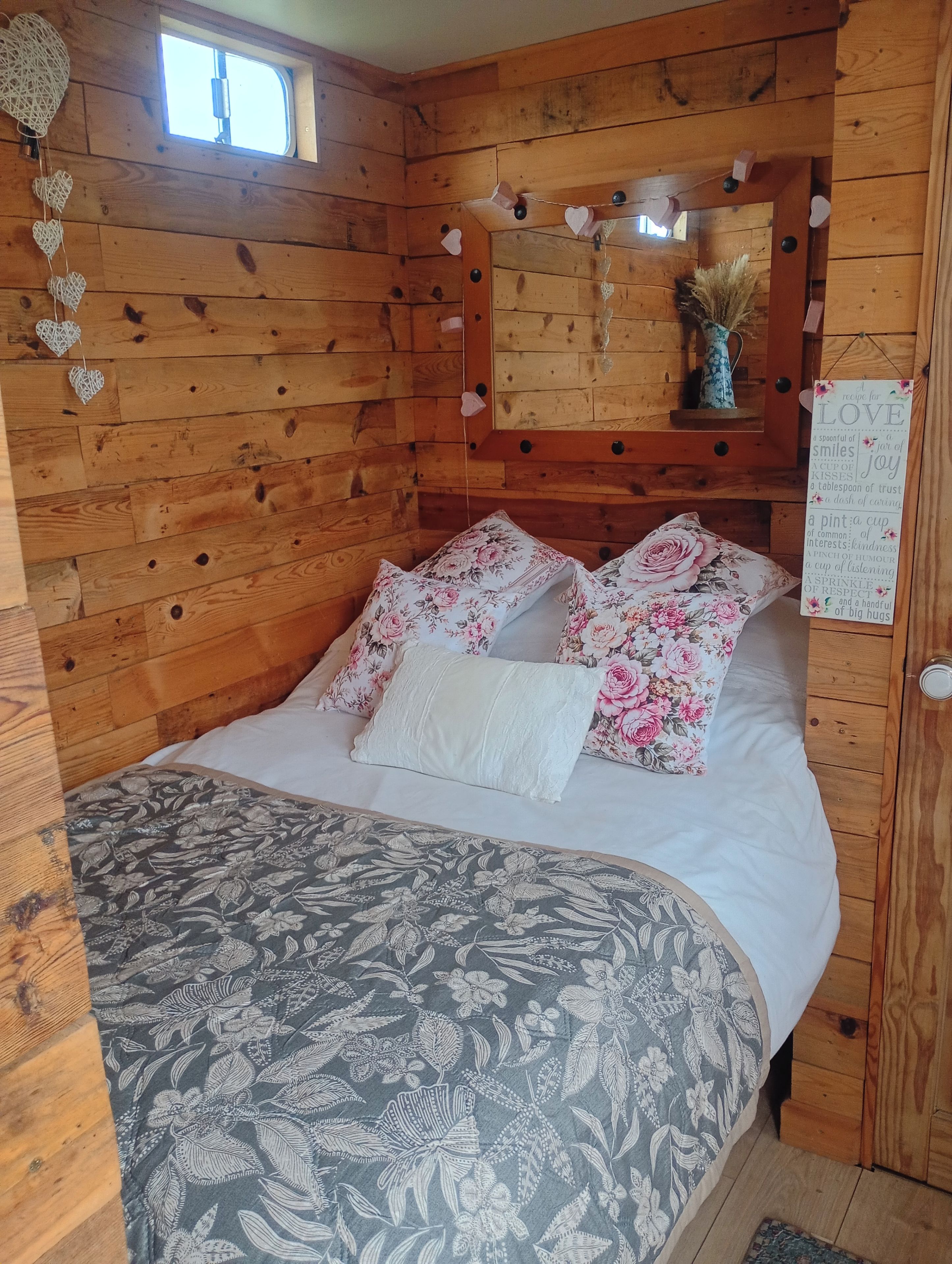 Ein Schlafzimmer einer Holzjagdhaus-ähnlichen Hütte mit einem Bett, blumigen Kissen, einer herzförmigen Dekoration und einem Schild mit dem Wort 'LOVE'.