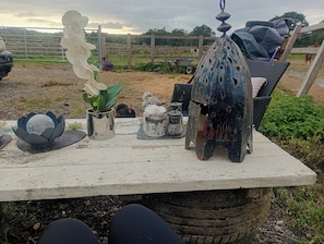 Cenário rústico ao ar livre com uma mesa de madeira exibindo diversos objetos decorativos, incluindo um comedouro de pássaros de metal, um vaso de metal e uma planta em vaso.