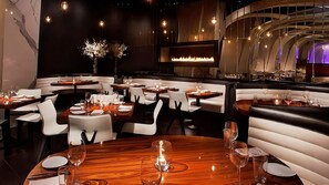 Ein Restaurantinneres mit runden Tischen, weißen Stühlen und einem zentralen Kamin.