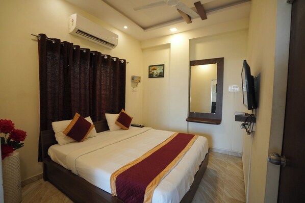 A hotel room with a bed, curtains, a wall-mounted TV, an air conditioning unit, and a painting.