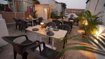 An outdoor dining area with tables and chairs, surrounded by potted plants and a building in the background.