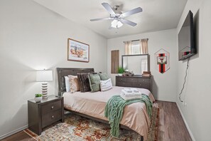 Une chambre à coucher comprenant un lit, une table de chevet, une commode, un ventilateur de plafond, des œuvres d’art, un miroir et un téléviseur fixé au mur.