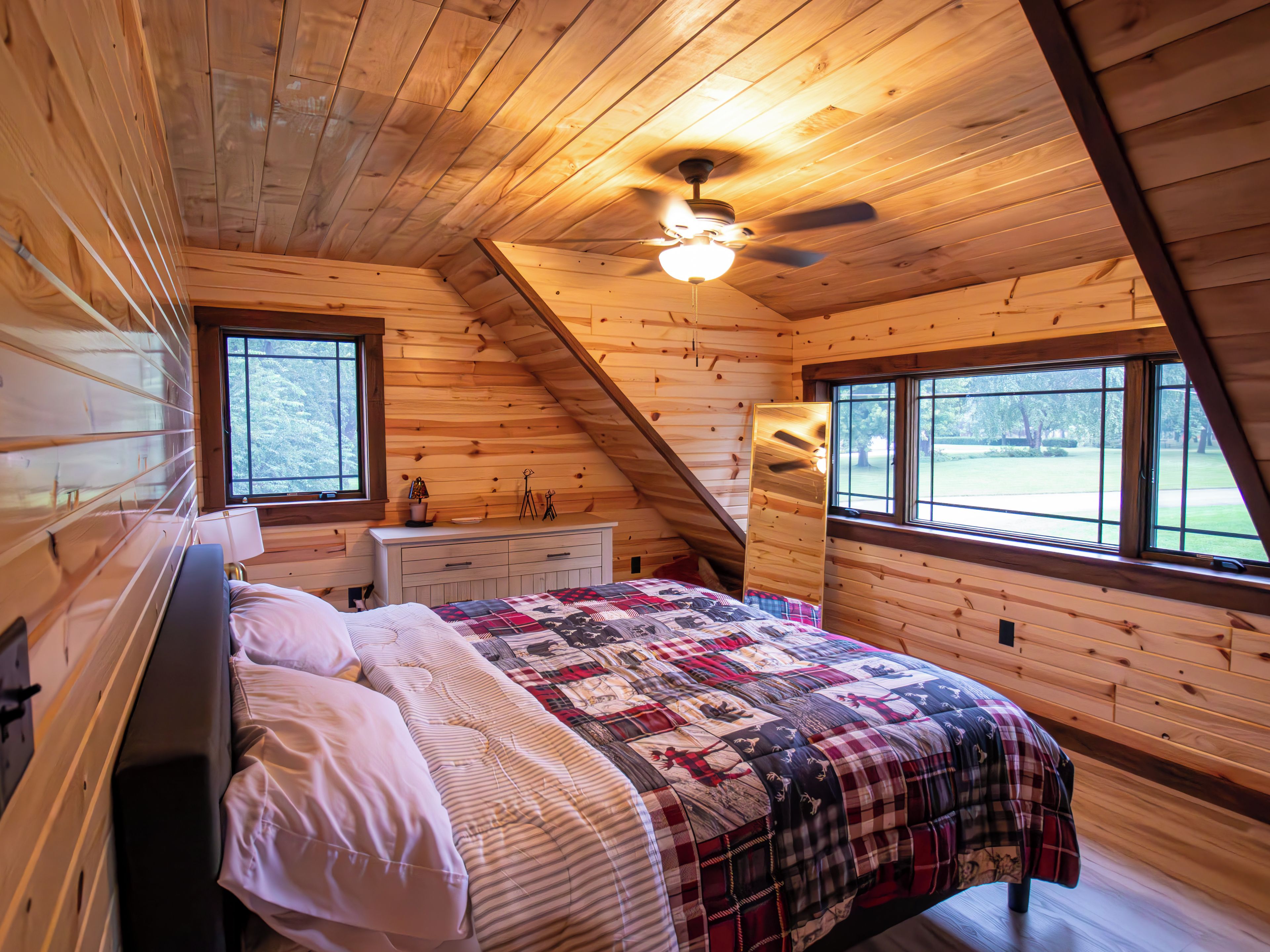 A bedroom with a bed, dresser, windows, and a sloped ceiling with a fan.