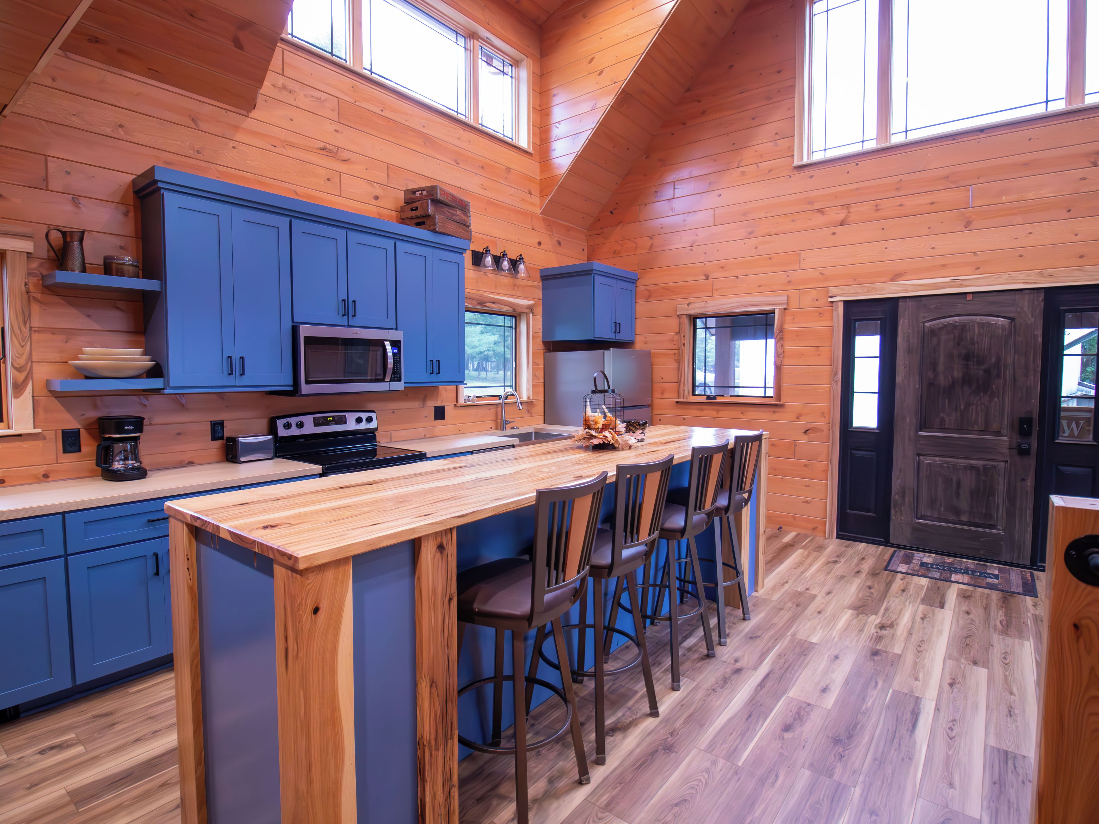A log cabin kitchen with blue cabinets, a central island with bar stools, and a large wooden door.
