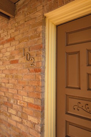 Un mur de briques portant le numéro « 102 » et une porte marron ornée d’un panneau décoratif.