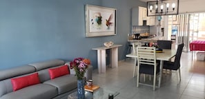 A modern living room with a grey sofa, red cushions, a coffee table with a vase of flowers, a dining area with a white table and chairs, and a kitchen in the background. - 4-bedroom townhome in lovely Puerto Morelos with AC and jacuzzi for 8 (Puerto Morelos)