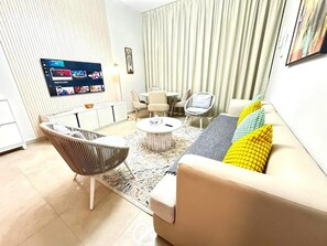 A modern living room with a flat-screen TV, a sofa with colorful cushions, a wicker chair, a round coffee table, and a dining area with a table and chairs. - 1BR Sparkle Tower Dubai Marina (Dubai)