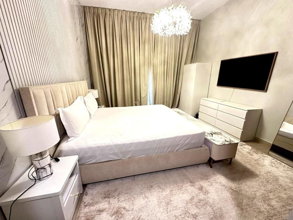 A modern bedroom with a large bed, a bedside table with a lamp, a dresser, a TV mounted on the wall, and a chandelier.