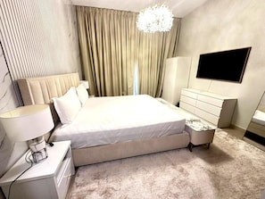 A modern bedroom with a large bed, a bedside table with a lamp, a dresser, a TV mounted on the wall, and a chandelier. - 1BR Sparkle Tower Dubai Marina (Dubai)