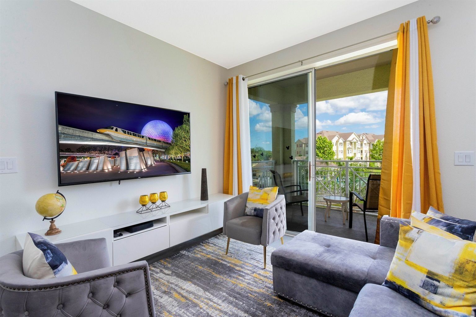 A modern living room with a flat-screen TV, a sofa, a chair, a small table, and a balcony with yellow curtains.