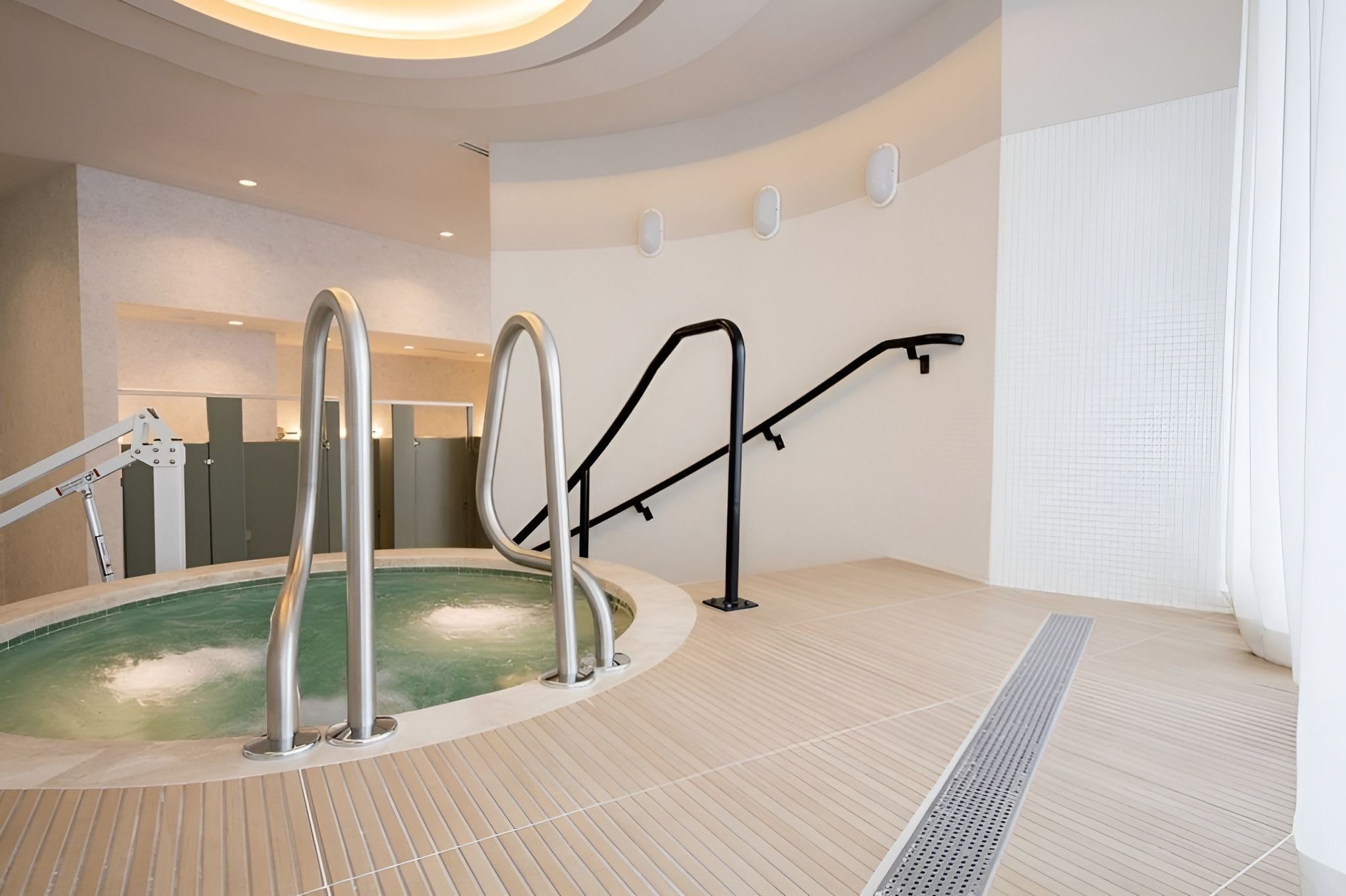 Un espace piscine intérieur moderne, doté d’un jacuzzi, de rampes en métal et d’un sol carrelé.
