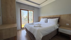 A bedroom with a large bed, wooden headboard, a nightstand, and a window with blinds.
