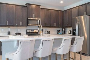 A modern kitchen with dark wood cabinets, stainless steel appliances, and a central island with bar stools. - Luxury 8BR Family Home Near Disney at Encore 54MA (Kissimmee)