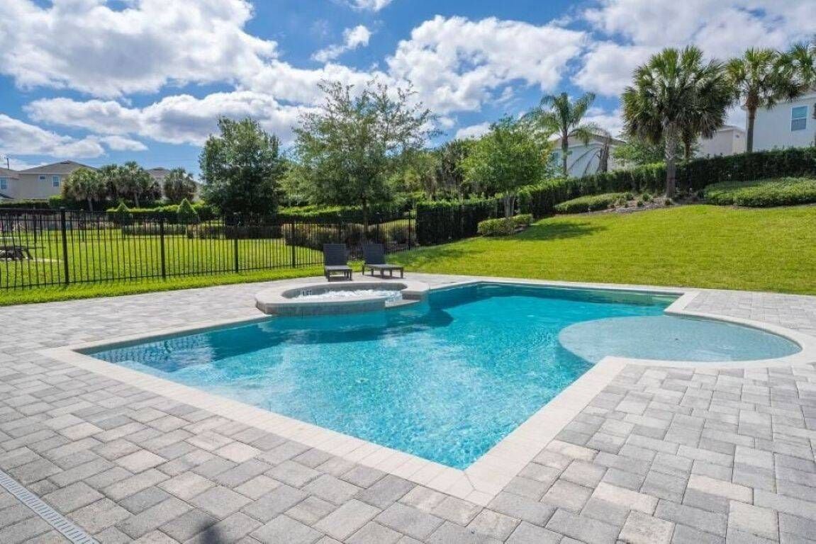 A swimming pool with a paved deck, surrounded by a fence and greenery.