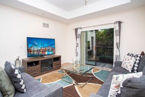 A living room with a flat-screen TV, a glass-top coffee table, grey sofas, and a patterned rug. - Modern Family Condo 15min From Disney (Kissimmee)