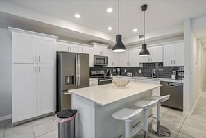 A modern kitchen with white cabinetry, stainless steel appliances, a central island with bar stools, and pendant lighting. - Cozy 2BR Condo 10 Min to Disney Pool (Kissimmee)