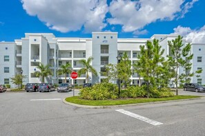 A modern multi-story building with a stop sign, parked cars, and a landscaped area with palm trees. - Cozy 2BR Condo 10 Min to Disney Pool (Kissimmee)