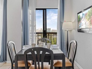 Een eetgedeelte met een tafel voor vier personen, stoelen, een aan de muur bevestigde televisie en een uitzicht op een stadsgezicht door een raam met gordijnen.