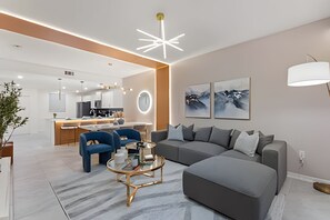Una sala moderna con un sofá seccional gris, un sillón azul y una mesa de centro de vidrio. Se ve una cocina con isla central e iluminación colgante al fondo.