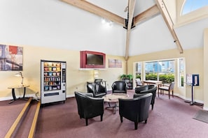 Ein Zimmer mit hoher Decke, ein Verkaufsautomat, mehrere schwarze Stühle, ein kleiner Tisch und ein an der Wand befestigter Fernseher.