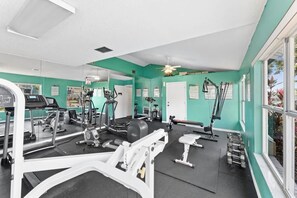 Ein Fitnessraum mit unterschiedlichen Trainingsgeräten, einem Deckenventilator und Blick ins Freie durch große Fenster.