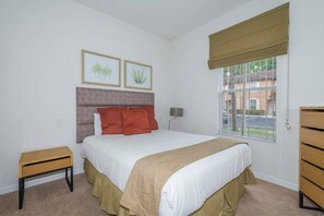 Quarto com cama, mesa de cabeceira, abajur, janela com persiana e duas gravuras botânicas emolduradas na parede.