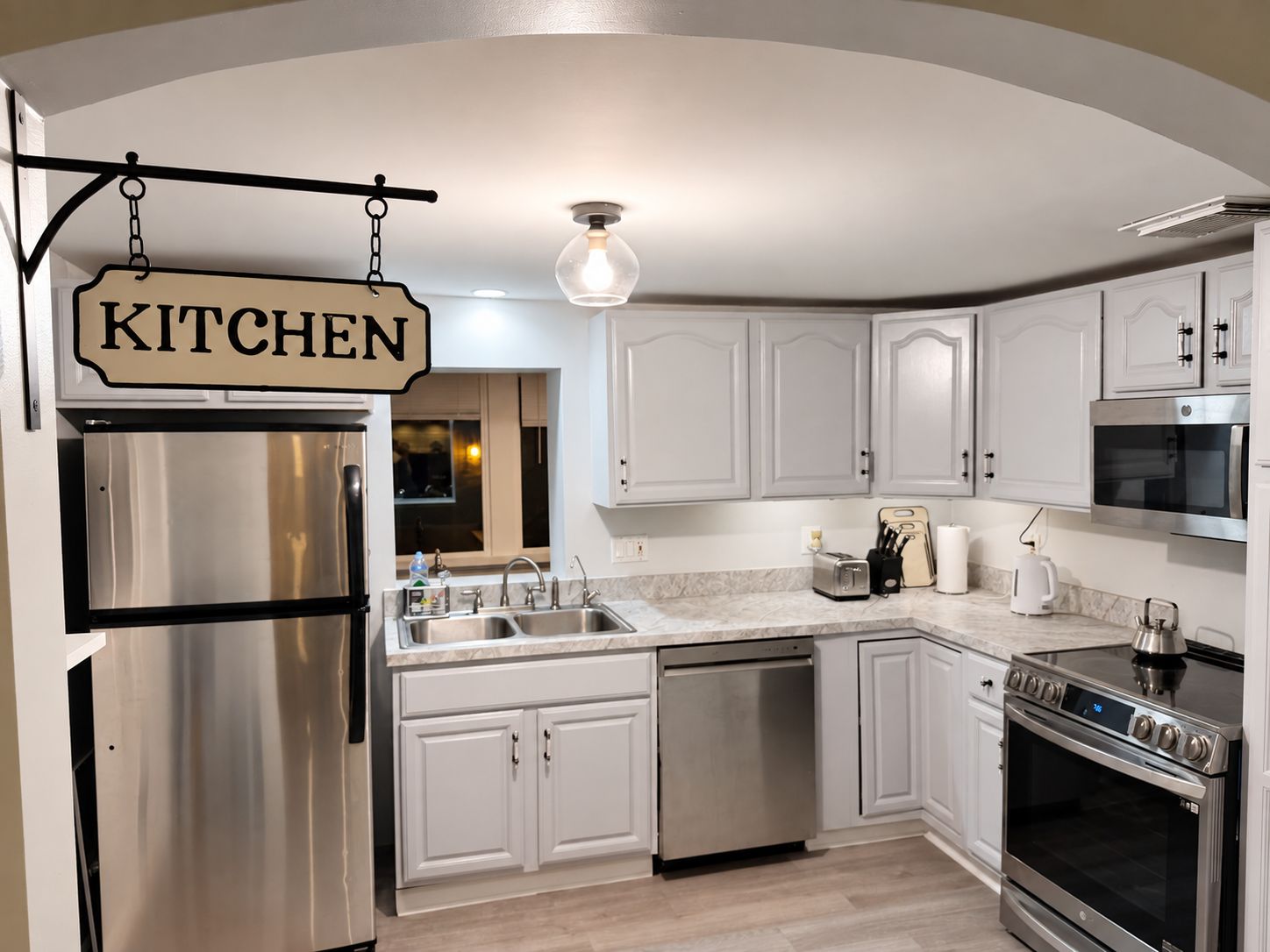 Una cocina moderna con electrodomésticos de acero inoxidable, armarios blancos y un letrero colgante que dice 'KITCHEN'.