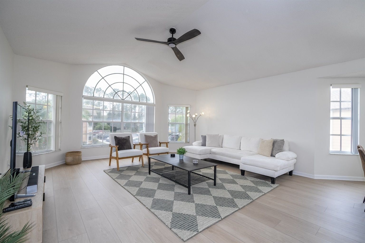 A spacious living room with a white sofa, two armchairs, a coffee table, and a ceiling fan. Large windows provide natural light and a view of the outside.