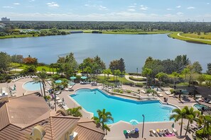 A large outdoor swimming pool surrounded by lounge chairs and umbrellas, with a view of a lake and distant buildings. - 3BR Vista Cay Condo Near Universal (Orlando)
