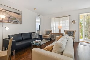 Ein modernes Wohnzimmer mit einer schwarzen Couch, beigen Sesseln und einem Glastisch. An der Wand hängt ein Gemälde und es gibt ein Fenster mit Jalousien.