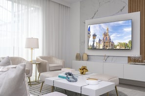 A modern living room with a large flat-screen TV displaying a castle, a sofa, a coffee table with books and decorative items, and a lamp. - 4BR Storey Lake Townhome, Private Pool (Kissimmee)