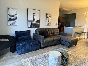 A modern living room with a grey sectional sofa, a blue armchair, and a patterned rug. There are three framed pictures on the wall, and a kitchen area is visible in the background. - Lake Buena Vista Resort - 2BR Condo (Orlando)