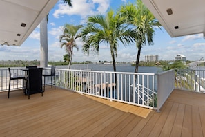 A wooden deck with a white railing, overlooking a body of water with palm trees and buildings in the distance. - Lured Away (Fort Myers Beach)