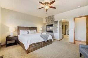 A bedroom with a wooden bed, two bedside tables with lamps, a stone accent wall, a fireplace, and a ceiling fan. - Timberline Hideout! Secluded Mtn Escape w Hot Tub (Woodland Park)