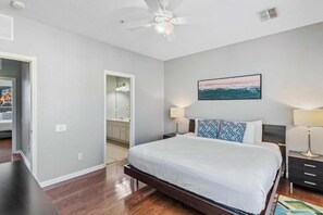 A bedroom with a large bed, a ceiling fan, a painting of a seascape, and a bathroom visible through an open door. - Modern 3BR Vista Cay Townhome (Orlando)