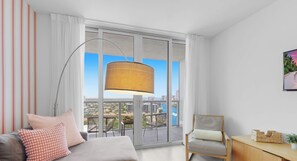 A modern living room with a sofa, a chair, a lamp, and a view of a cityscape through large windows. - BW APT 2BR Resort Style Suite - Bay & City Views (Hallandale Beach)