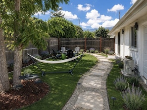 A backyard with a hammock, a patio table, and chairs, surrounded by a wooden fence and a house. - Pixie and Orchard Cottages - Sleeps 7 (Ojai)