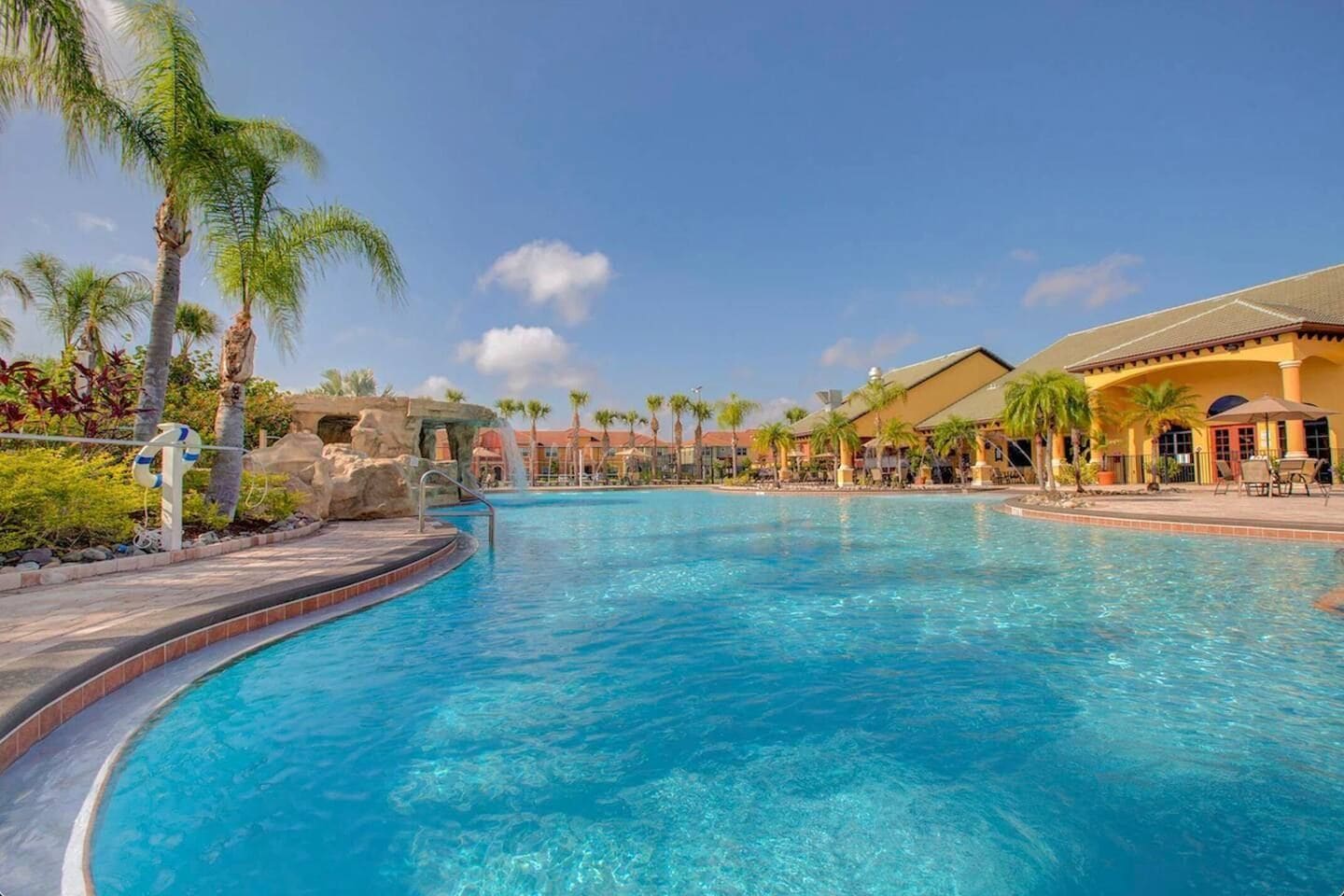 A large outdoor swimming pool with clear blue water, surrounded by palm trees and a resort building with a tiled roof.