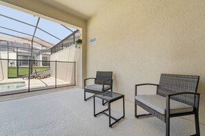A covered outdoor seating area with two chairs and a small table, overlooking a swimming pool and a grassy lawn. - Championsgate Pool Home Near Disney Parks (Davenport)