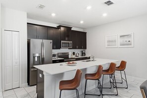 A modern kitchen with dark cabinets, stainless steel appliances, a central island with bar stools, and a white countertop. - Championsgate Pool Home Near Disney Parks (Davenport)