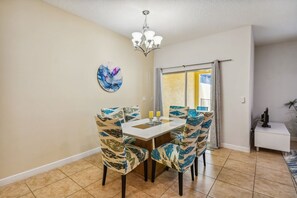 A dining area with a table and chairs, a chandelier, a painting on the wall, and a view through a window with curtains. - 4BR Paradise Palms Townhome, Heated Pool (Kissimmee)