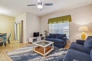 A living room with a blue sofa, a glass coffee table, a TV, and a ceiling fan. - 4BR Paradise Palms Townhome, Heated Pool (Kissimmee)