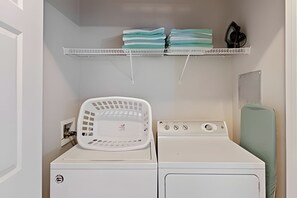 A laundry area with a washing machine, a dryer, a white laundry basket, and a shelf with folded clothes and a black object. - Spacious 3BR Condo at Vista Cay Resort (Orlando)