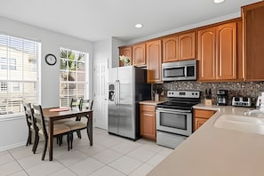 A modern kitchen with wooden cabinets, stainless steel appliances, and a dining area with a table and chairs. - Vista Cay 3BR Townhome Near Universal & Occc (Orlando)