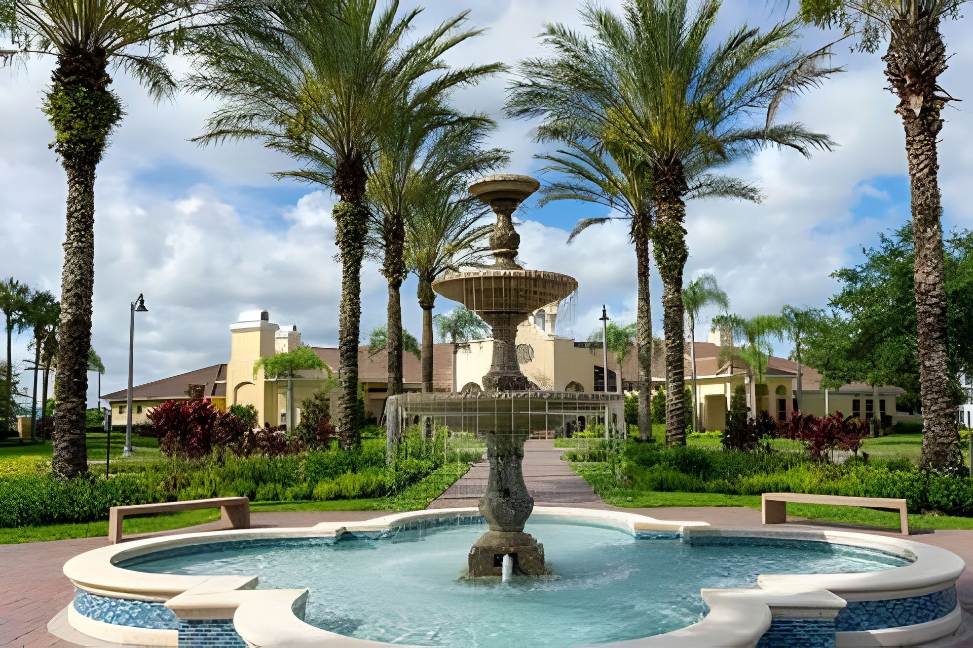 A fountain with a tiered design, surrounded by palm trees and a pool, with a building in the background.