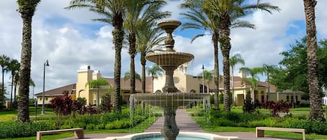 A fountain with a tiered design, surrounded by palm trees and a pool, with a building in the background.