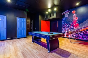 A billiards table in a room with wooden flooring, a large screen displaying a basketball game, and blue doors. - Windsor Hills Luxe 4BR Villa w/ Pool & Game Room (Kissimmee)