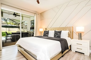 A modern bedroom with a large bed, two bedside lamps, a nightstand, and a sliding glass door leading to an outdoor patio with a table and chairs. - Windsor Hills Luxe 4BR Villa w/ Pool & Game Room (Kissimmee)